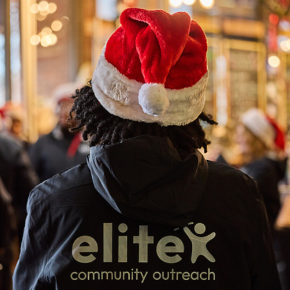 A child in a Santa hat and Elite Community Outreach hoodie faces away from the camera looking at members in the choir.