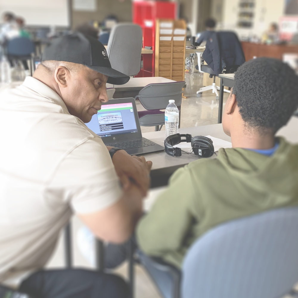 Carl Cannon provides guidance to a Game Changer Safe School student. He is squatting down next to a seated student who is looking into the screen of his laptop.